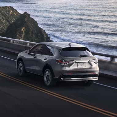 2027 Honda HR-V EX-L in Modern Steel Metallic rear view driving on a coastal road near the ocean (Subcompact Crossover SUV).